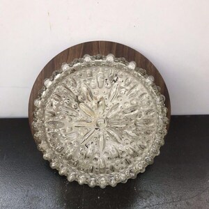 Puede incluir: Una lámpara de techo vintage. La lámpara tiene una pantalla de vidrio transparente y texturizado con un diseño de estrella, rodeada por un borde con cuentas. Está montada sobre una base redonda de madera marrón oscuro. La lámpara está diseñada para montarse en el techo.