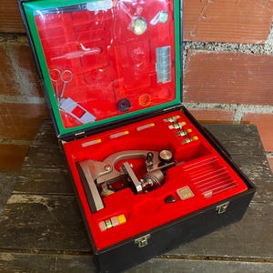 May include: A black case with a red interior, holding a silver microscope and scientific equipment. The case has a green trim and is on a wooden surface. The tools include vials, slides, and other small instruments.