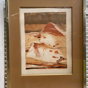 May include: A framed abstract print with a brown, orange, and white color scheme. The print depicts a landscape with hills and valleys. The artist's signature is visible in the bottom right corner.