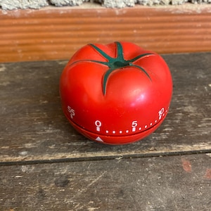 May include: A red tomato-shaped kitchen timer with a green stem. The timer has white numbers and markings around the base, indicating minutes. It sits on a weathered wooden surface.