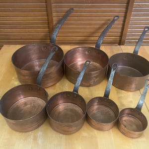 May include: A set of six graduated copper saucepans with dark, riveted iron handles. The pots feature a polished metallic copper exterior with visible surface textures and rounded sides, arranged in two rows on a flat wooden surface.