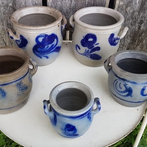 May include: Five blue and white ceramic crocks with handles. The crocks are decorated with blue floral and geometric patterns. The crocks are arranged on a white table with a wooden background.