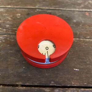 May include: A red, circular gauge with a white dial and a needle pointing to zero. The gauge has a blue accent and is set against a weathered wooden surface. The dial displays numbers from 0 to 5.