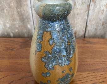 Könnte beinhalten: Eine kleine, handgefertigte Keramikvase mit einer einzigartigen kristallinen Glasur. Die Vase hat einen abgerundeten Körper und einen schmalen Hals. Die Glasur wechselt von einem warmen, goldbraunen Farbton am Boden zu einem kühlen Blau mit kristallinen Mustern und einem hellgrünen Rand.