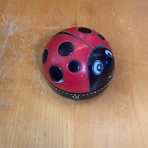 May include: A red and black ladybug-shaped kitchen timer with a rounded top featuring black spots. The base is black with white markings, including the number 10, and a rotating mechanism for setting time intervals for cooking or other tasks.