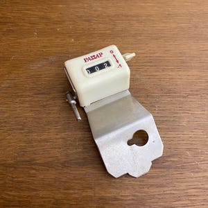 May include: White counter with a silver metal bracket. The counter has a red label that says "PASSAP" and displays the number "002".