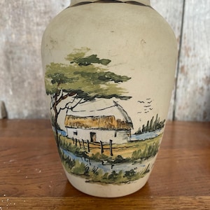 Peut inclure: Un vase en céramique de couleur crème avec une scène peinte à la main d'un chalet avec un toit de chaume, un arbre et des oiseaux volant au-dessus d'une rivière. Le vase a une bordure brune.