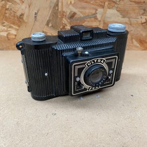 May include: A black vintage Ultra Spec camera with a silver lens and a silver knob on the top. The camera has a black and white design with the words "Ultra Spec" printed on the front.