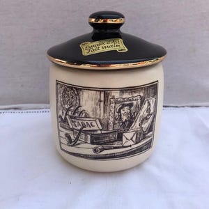May include: A cream-coloured ceramic tobacco jar with a black lid and gold trim. The jar has a black and white illustration of a man, tobacco, and a pipe. The lid has the text "Emaux d'Art Fait main".