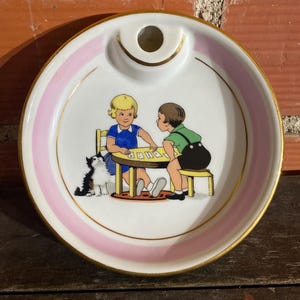 May include: A white ceramic ashtray with a pink stripe and gold trim. The centre features a painted scene of two children playing cards at a table, with a black and white cat. The ashtray has a small circular indentation.