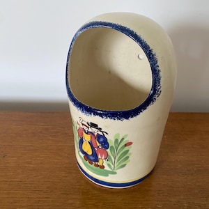 Puede incluir: Un macetero de pared de cerámica blanca con un borde azul y una escena pintada de dos personas con ropa tradicional. El macetero tiene un agujero en la parte posterior para colgarlo.