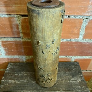 May include: A cylindrical wooden vase with a dark brown, circular top. The vase is a light brown colour with dark brown accents and several small, decorative floral embellishments. The vase sits on a weathered wooden surface against a brick wall.