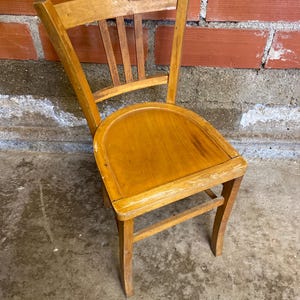 Peut inclure: Petite chaise en bois vieilli, avec une assise incurvée et un dossier à lattes. Le bois clair présente des signes d'usure, avec des zones décolorées et des rayures. La chaise est placée devant un mur de briques.