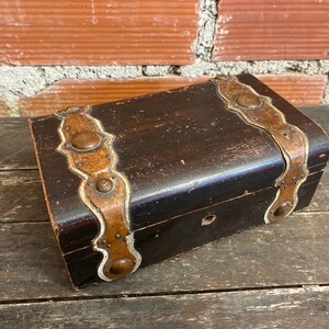 Puede incluir: Pequeña caja rectangular de madera con acabado marrón oscuro. Destacan herrajes metálicos decorativos en tono cobre. La caja cerrada descansa sobre una superficie de madera envejecida, sugiriendo un artículo antiguo o vintage.