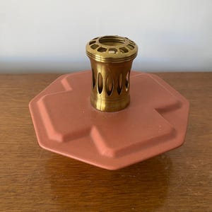 May include: A vintage oil lamp burner with a brass top and a terracotta-colored base. The brass top has a series of oval-shaped openings. The base is a unique, geometric shape. The lamp is designed to hold oil and provide light.