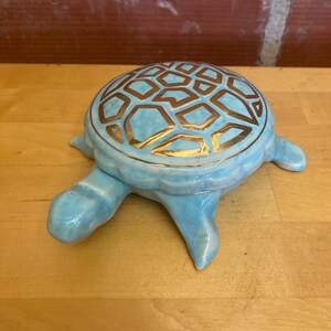 Könnte beinhalten: Hellblaue Keramik-Schildkrötenfigur mit goldenem geometrischen Muster auf dem Panzer. Die Schildkröte hat vier Beine, einen Kopf und einen Schwanz. Die Figur steht auf einer Holzoberfläche.