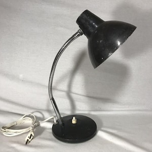 Puede incluir: Una lámpara de escritorio negra con un cuello flexible plateado y una base redonda. La lámpara tiene una pantalla en forma de cúpula y un cable de alimentación blanco. La lámpara está diseñada para usar en un escritorio o mesa.
