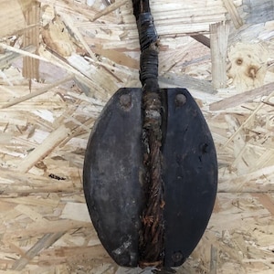 Peut inclure: Une poulie en bois antique avec un corps ovale foncé et une corde épaisse et usée traversant son centre. La corde est enveloppée d'un matériau sombre. La poulie est posée sur un fond en bois clair et texturé.