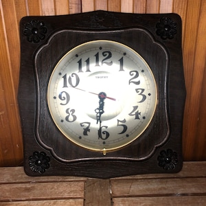 Può includere: Un orologio con cornice in legno scuro con una cornice dorata e un quadrante bianco. Il quadrante dell'orologio ha numeri romani neri e la parola "Trophy" stampata su di esso.