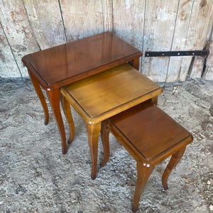 May include: A set of three nested tables made of polished wood with a smooth finish. Each table features a rectangular top with rounded corners and four curved legs that extend outward, allowing the smaller tables to slide under the largest one.