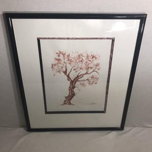 Op de afbeelding: Ingelijste aquarel schilderij van een boom met roodbruine stam en takken, en lichtroze bladeren. Het kunstwerk staat tegen een witte achtergrond met een getextureerde rand, omlijst door een zwart frame.