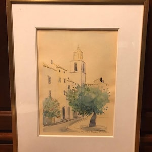 May include: Watercolor painting of a European village scene, framed in gold. The artwork depicts buildings, a church tower, and a tree along a street. The painting is set against a light beige background, with a white mat.