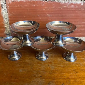 Puede incluir: Un juego de cinco pequeños cuencos de postre plateados y brillantes. Cada cuenco tiene un cuenco redondo y poco profundo sobre una base corta con pedestal. Los cuencos están colocados sobre una superficie de madera, con una pared de ladrillo al fondo.
