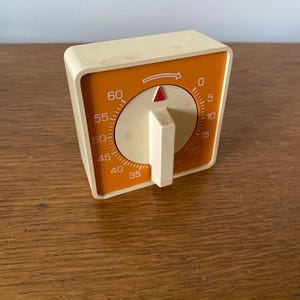 May include: A vintage kitchen timer with a cream-colored body and an orange face. The timer features a dial with numbers from 0 to 60, a red indicator, and a cream-colored knob. The timer is likely from the mid-20th century.