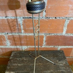 Peut inclure: Cendrier vintage avec un motif noir tressé et un bord en métal argenté. Le cendrier est posé sur un support long et fin avec des volutes décoratives et une base à trois pieds. Le support est en métal.