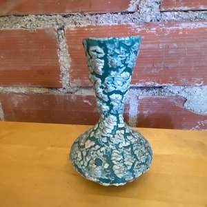 May include: A small vase with a bulbous base and a flared rim, featuring a textured surface with a crackled pattern in shades of deep teal green and off-white. The object has a distinct hourglass shape and a matte finish across its entire exterior.