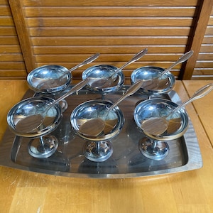 Könnte beinhalten: Ein Set aus sechs silberfarbenen Dessertschalen aus Metall mit passenden Löffeln, angeordnet auf einem rechteckigen silberfarbenen Serviertablett. Jede Schale hat eine breite, flache Form auf einem Standfuß und dazu passende Löffel.
