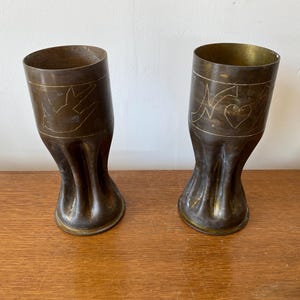 Pode incluir: Duas xícaras de latão antigas com desenhos decorativos. As xícaras são feitas de projéteis de artilharia reutilizados e têm um aspecto rústico e vintage.