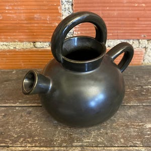 Puede incluir: Una tetera de cerámica gris oscuro con cuerpo redondeado, asa curva y boquilla. La tetera está sobre una superficie de madera. El asa es arqueada y la boquilla es corta e inclinada. La tetera tiene un acabado mate.