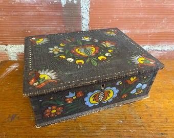 May include: A dark brown wooden box with a hinged lid, decorated with colourful floral folk art. The box has a keyhole and is painted with red, yellow, blue, and green flowers and leaves. The box is on a wooden surface.