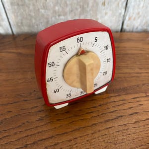 Puede incluir: Un temporizador de cocina rojo vintage con una esfera blanca y números negros. El temporizador tiene una perilla beige y pies blancos. El temporizador está configurado para aproximadamente 30 minutos. Es una herramienta de cocina retro.