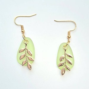 May include: A pair of earrings with pale green, teardrop-shaped pendants and gold-coloured leaf designs. The earrings have gold-coloured hooks. The background is white.