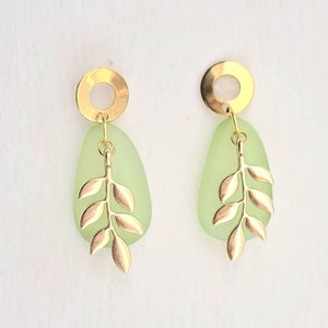 May include: Gold-coloured earrings with a leaf design. Each earring has a light green, teardrop-shaped stone, a gold circle at the top, and a gold leaf pendant. The earrings are set against a white background.