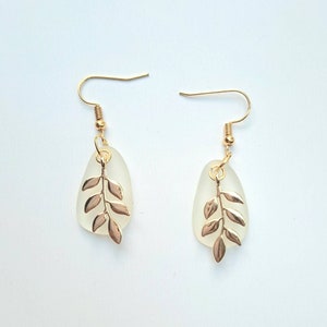May include: A pair of gold-coloured earrings with a leaf design. Each earring features a teardrop-shaped, frosted glass pendant with a gold-coloured leaf charm. The earrings have gold-coloured ear wires. The background is a solid white.