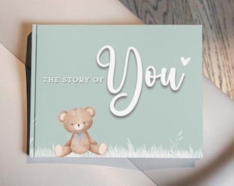 May include: A baby book with a light teal cover featuring the text "THE STORY OF You" in white script. A watercolor teddy bear with a blue bow sits below the text. The book is on a white surface.