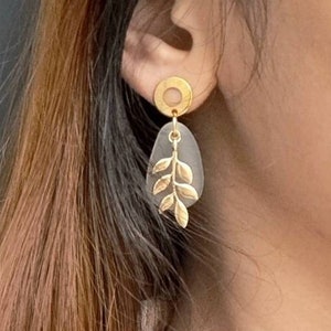 May include: Gold-coloured earrings with a leaf design. Each earring has a gold circle stud, a grey teardrop-shaped element, and a gold leaf accent. These earrings are a stylish accessory.