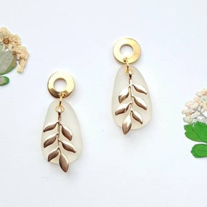 May include: Gold-coloured earrings featuring a leaf design. Each earring has a gold circle, a white oval centre, and a gold leaf motif. Dried flowers are in the background, set against a white surface.