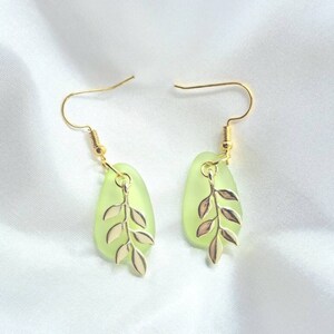 May include: A pair of gold-coloured hook earrings. Each earring features a light green, teardrop-shaped charm with a gold-coloured leaf design. The earrings are displayed on a white, textured surface.