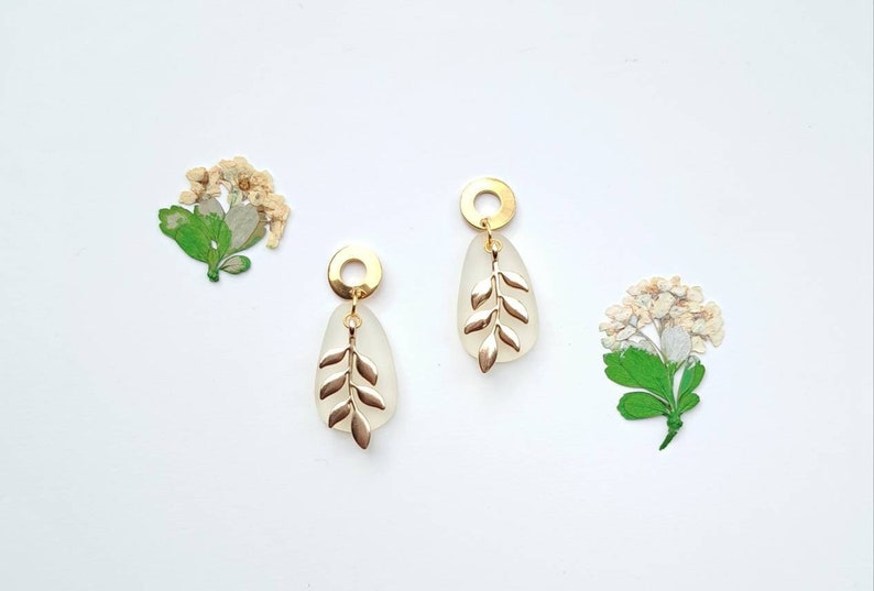 May include: Gold-coloured earrings featuring a leaf design. Each earring has a gold circle, a white oval centre, and a gold leaf motif. Dried flowers are in the background, set against a white surface.