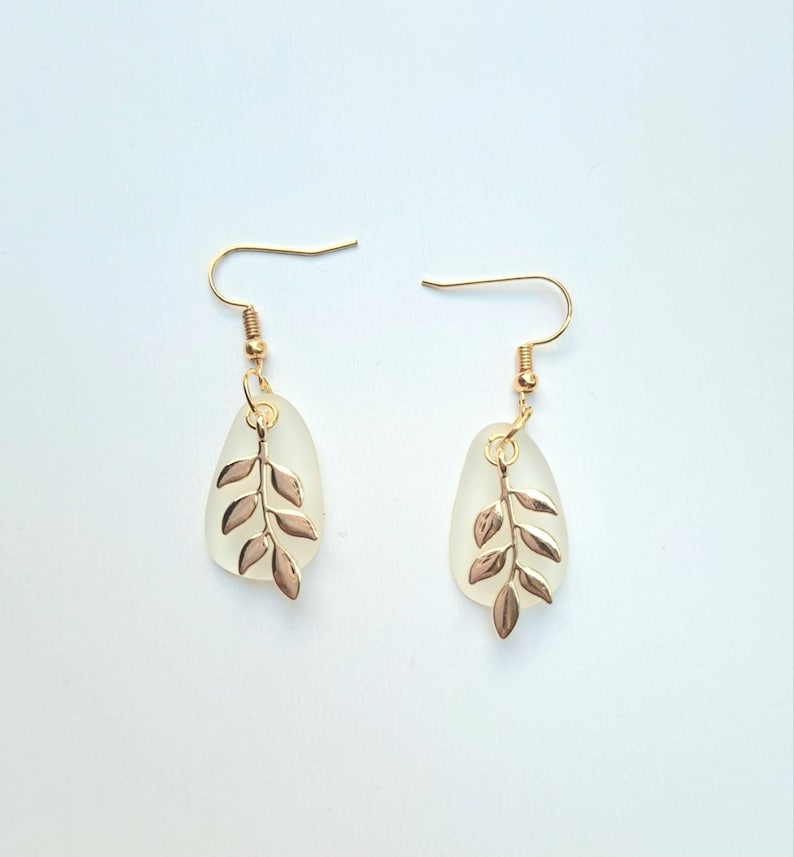 May include: A pair of gold-coloured earrings with a leaf design. Each earring features a teardrop-shaped, frosted glass pendant with a gold-coloured leaf charm. The earrings have gold-coloured ear wires. The background is a solid white.