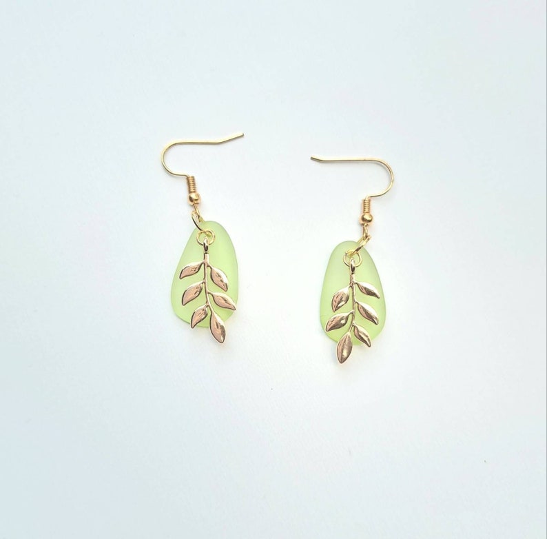 May include: A pair of earrings with pale green, teardrop-shaped pendants and gold-coloured leaf designs. The earrings have gold-coloured hooks. The background is white.