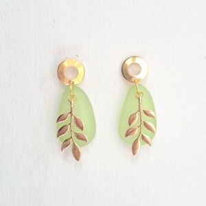 May include: A pair of gold-coloured earrings featuring a light green, teardrop-shaped stone and a gold-coloured leaf design. The earrings have a circular gold-coloured top. Set against a white background.