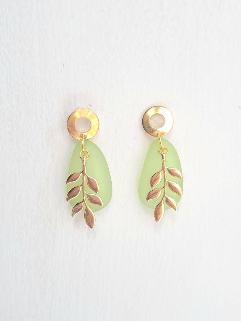 May include: A pair of gold-coloured earrings featuring a light green, teardrop-shaped stone and a gold-coloured leaf design. The earrings have a circular gold-coloured top. Set against a white background.