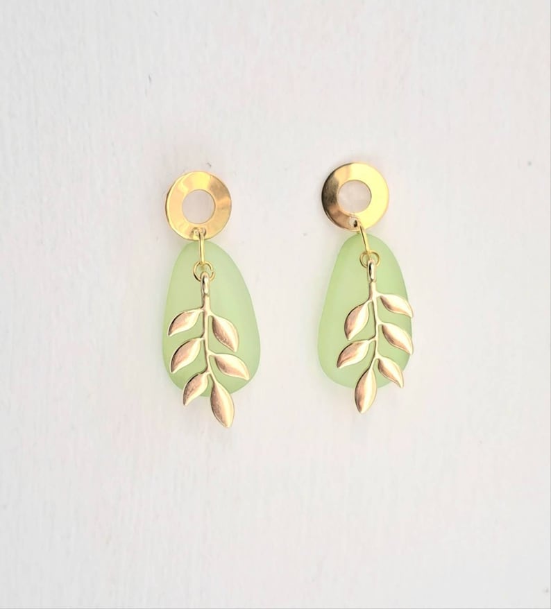 May include: Gold-coloured earrings with a leaf design. Each earring has a light green, teardrop-shaped stone, a gold circle at the top, and a gold leaf pendant. The earrings are set against a white background.
