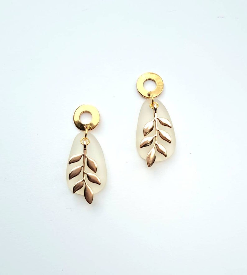 May include: A pair of gold-coloured earrings. Each earring has a gold-coloured circular top, a frosted teardrop-shaped pendant, and a gold-coloured leaf design. The earrings are displayed against a white background.