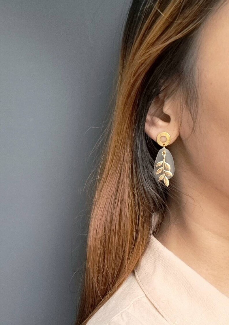 May include: Gold-coloured earrings with a leaf design. Each earring has a gold circle stud, a grey teardrop-shaped element, and a gold leaf accent. These earrings are a stylish accessory.